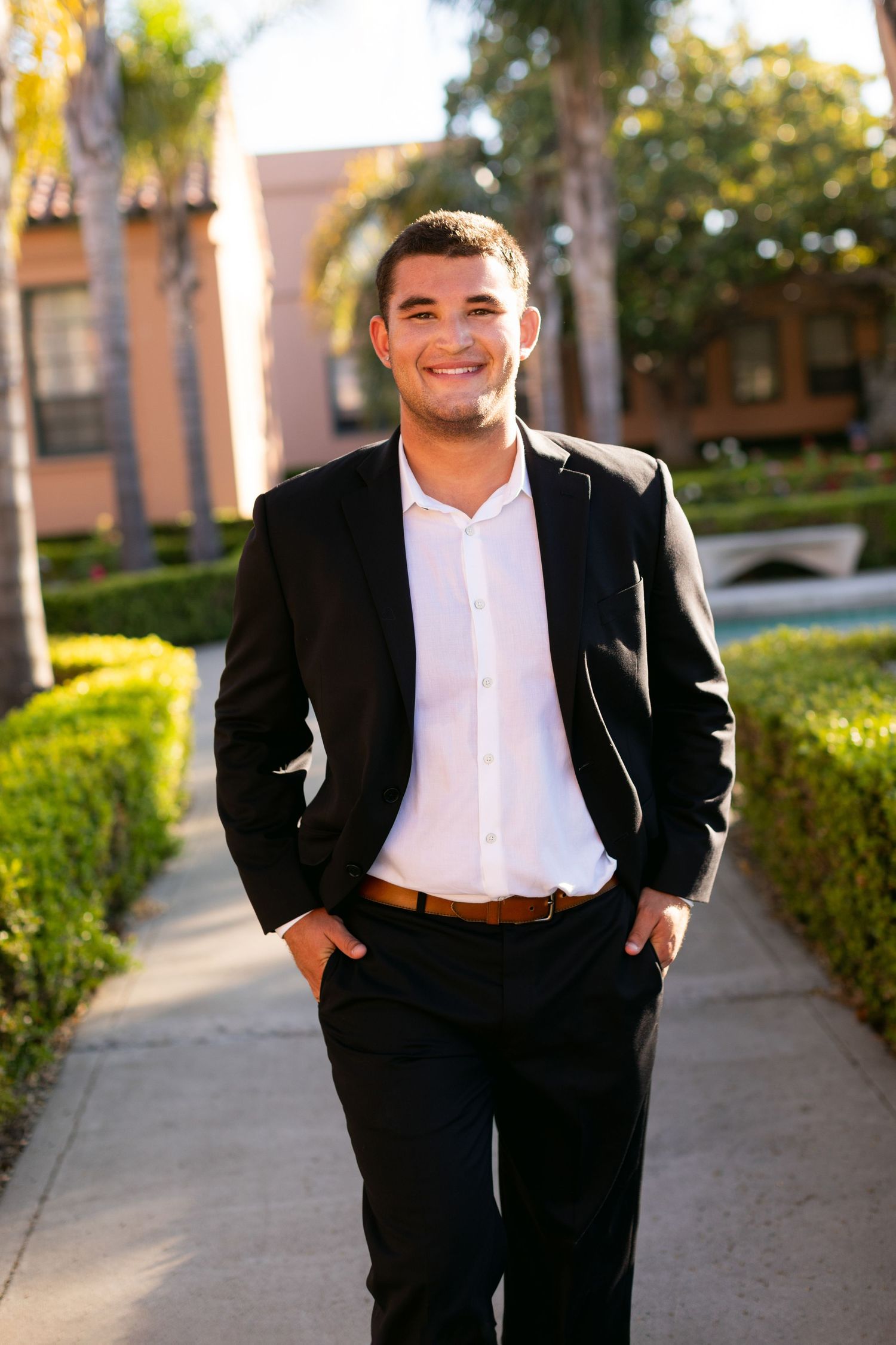 Professional portrait showing someone in a black suit and white shirt standing outdoors on a pathway with palm trees.