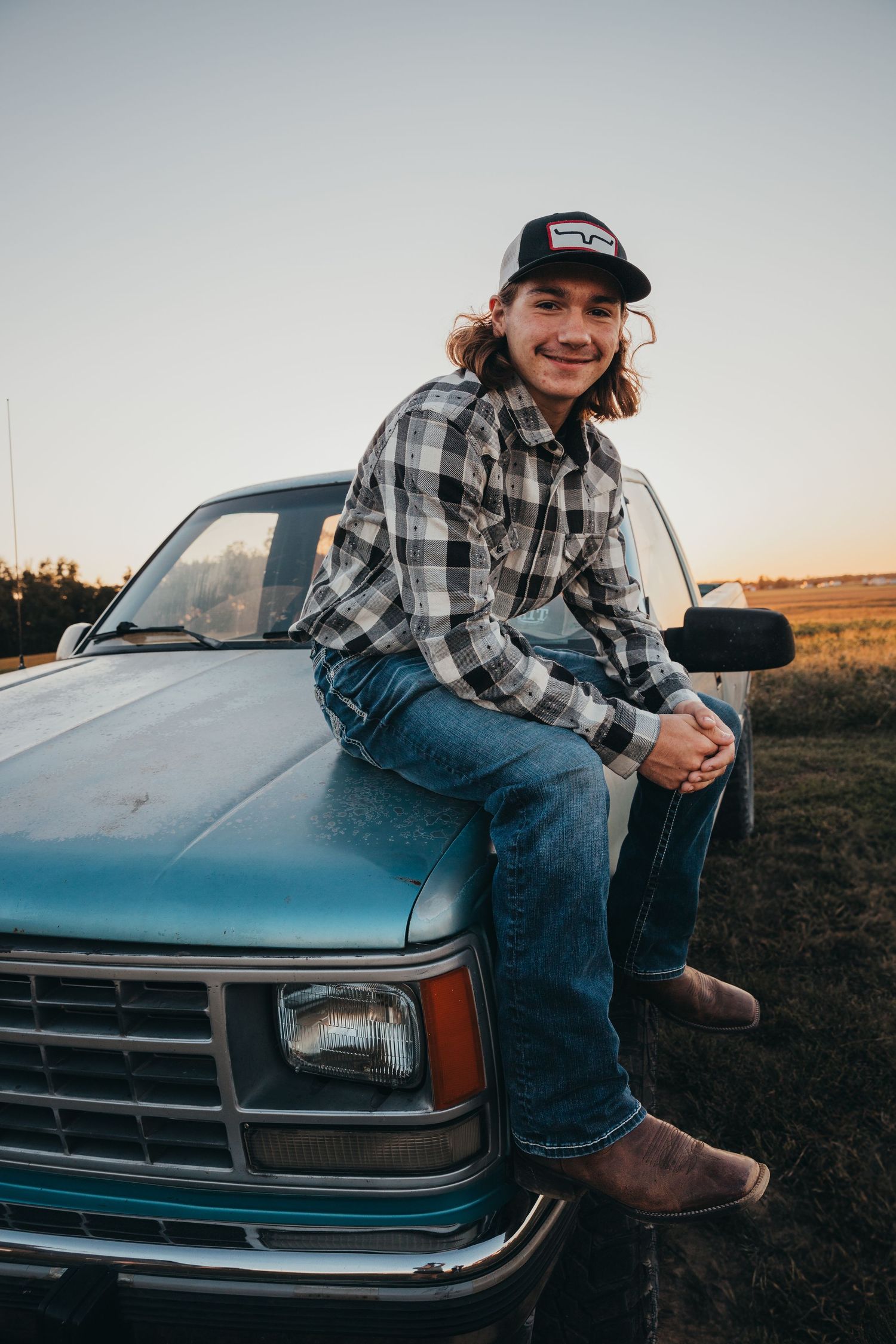 Person in plaid shirt and baseball cap sits on hood of vintage blue pickup truck in rural setting at sunset.