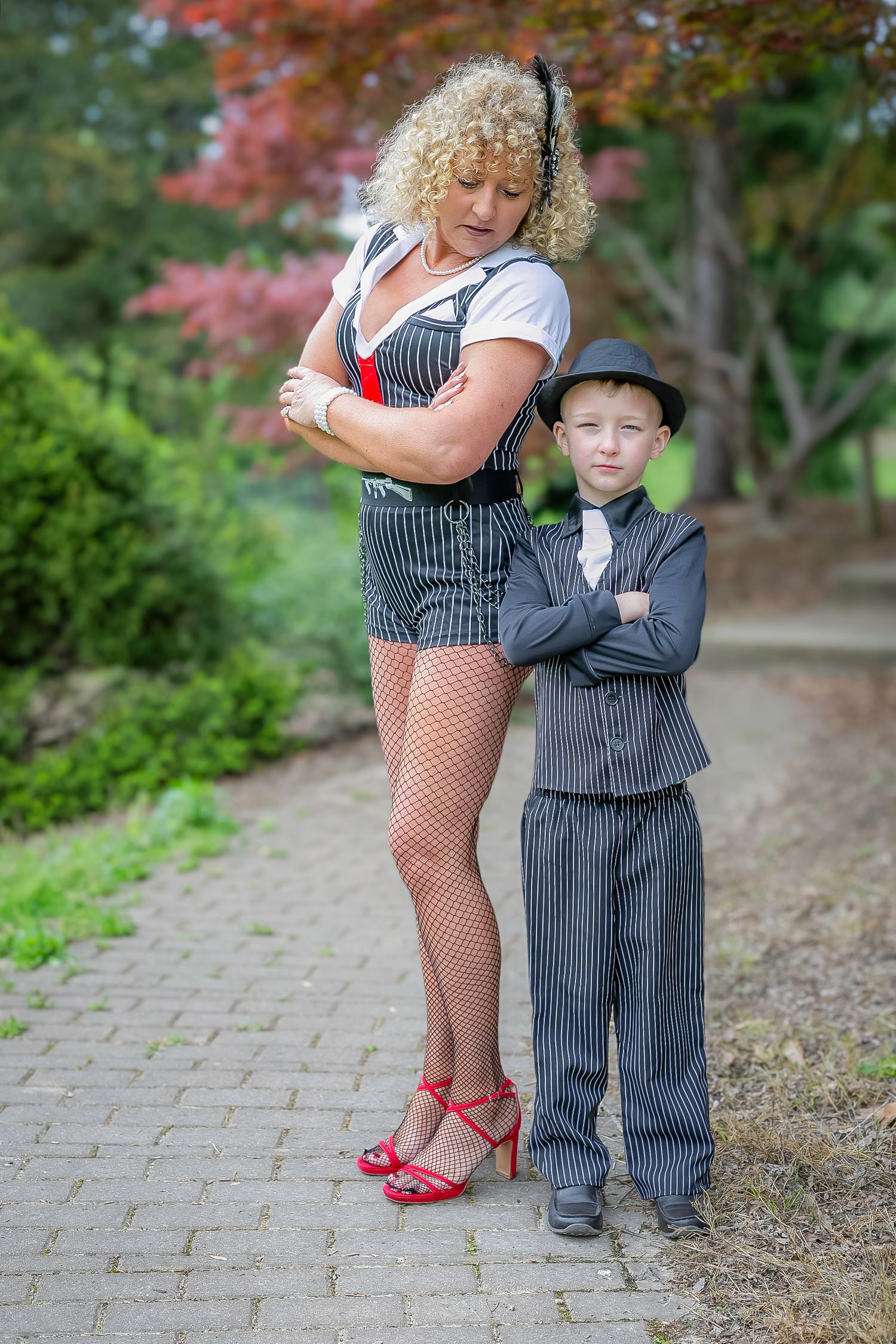 Two performers in pinstripe outfits strike stylized poses on a garden path with red foliage in the background.