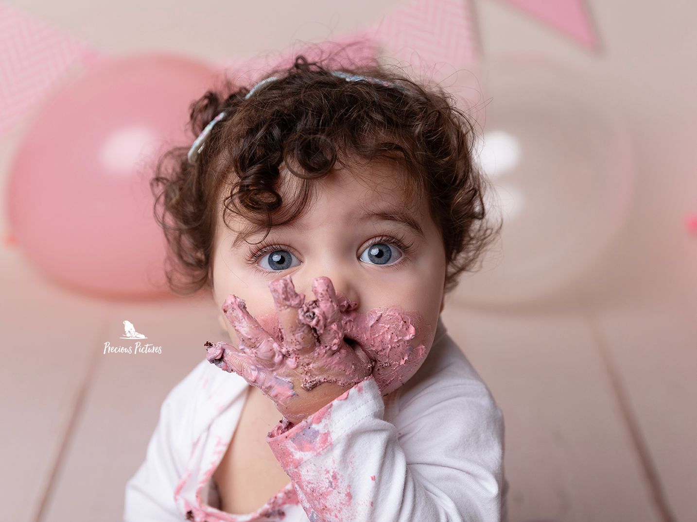 Close-up of a curly-haired baby girl with pink frosting on her face and hands, professional cake smash photography by Precious Pictures