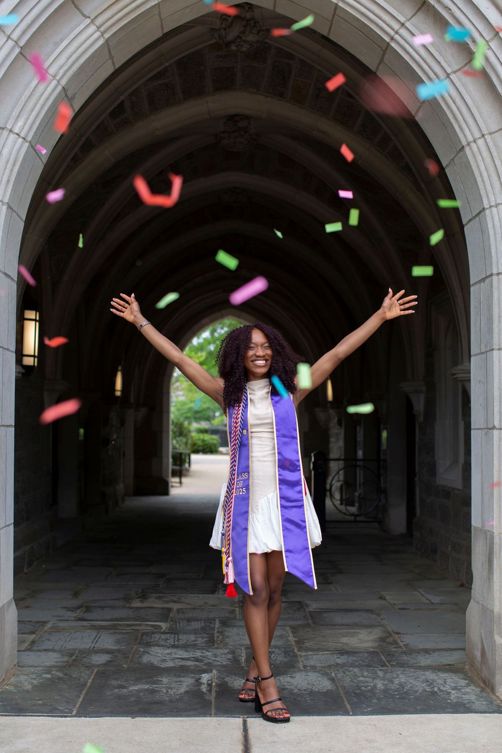 Joyful graduate in white dress and purple stole celebrates with colorful confetti under stone archway.