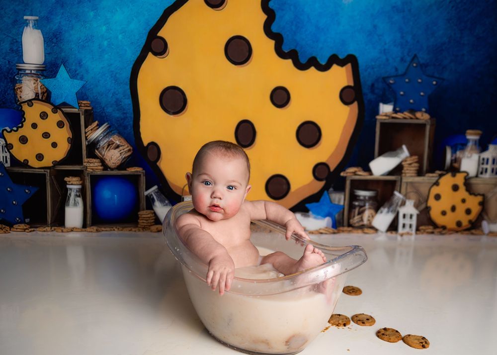 A baby sits in a vintage bathtub with a large yellow cookie backdrop and scattered cookie decorations in a studio setting.