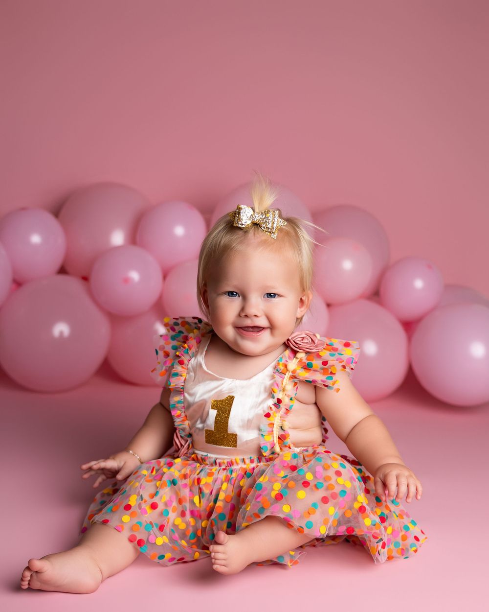 Baby girl in colorful polka dot dress celebrating her 1st birthday with pink balloons backdrop.