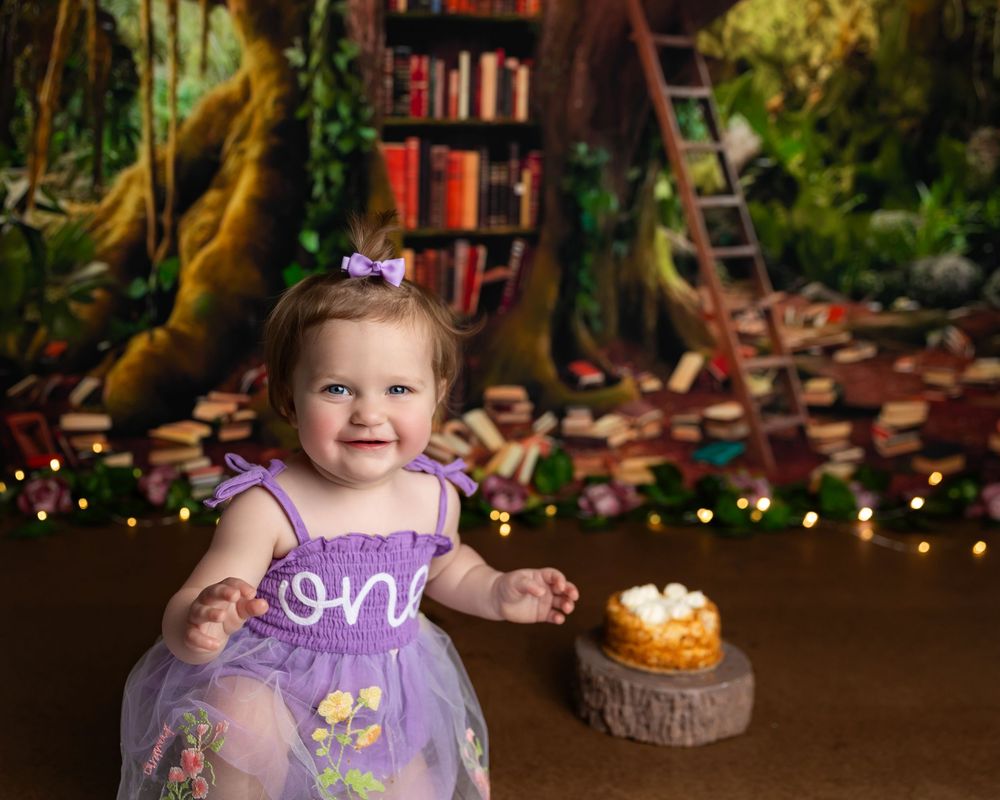 Smiling baby girl in purple dress celebrates first birthday with cake smash against enchanted library backdrop.