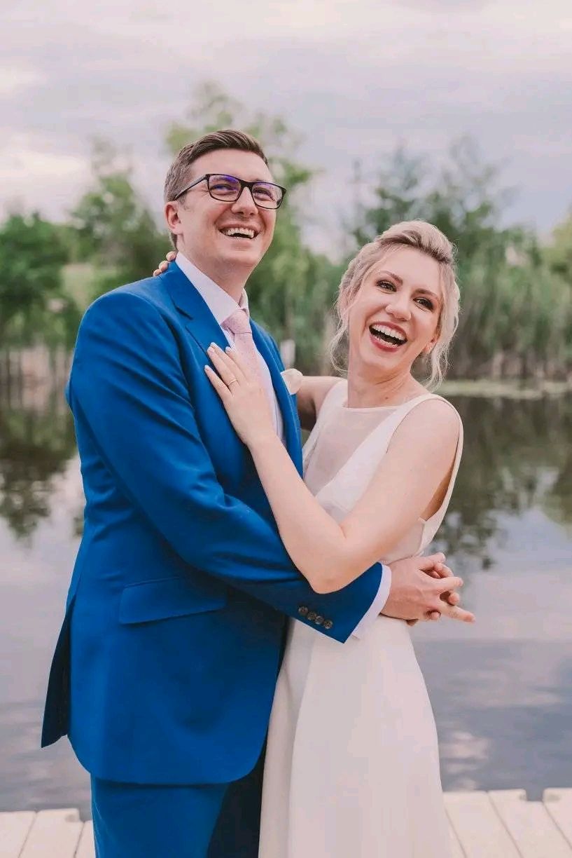 A joyful couple in blue suit and white dress share candid laughs during their outdoor wedding photoshoot by the water.