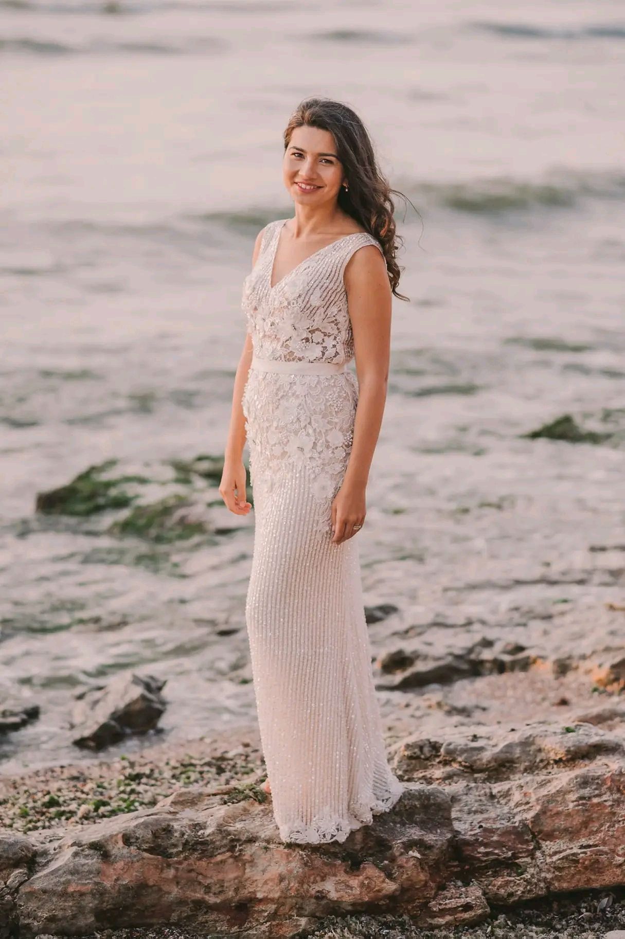 A person in a beaded cream-colored dress poses on rocky beach terrain at sunset.