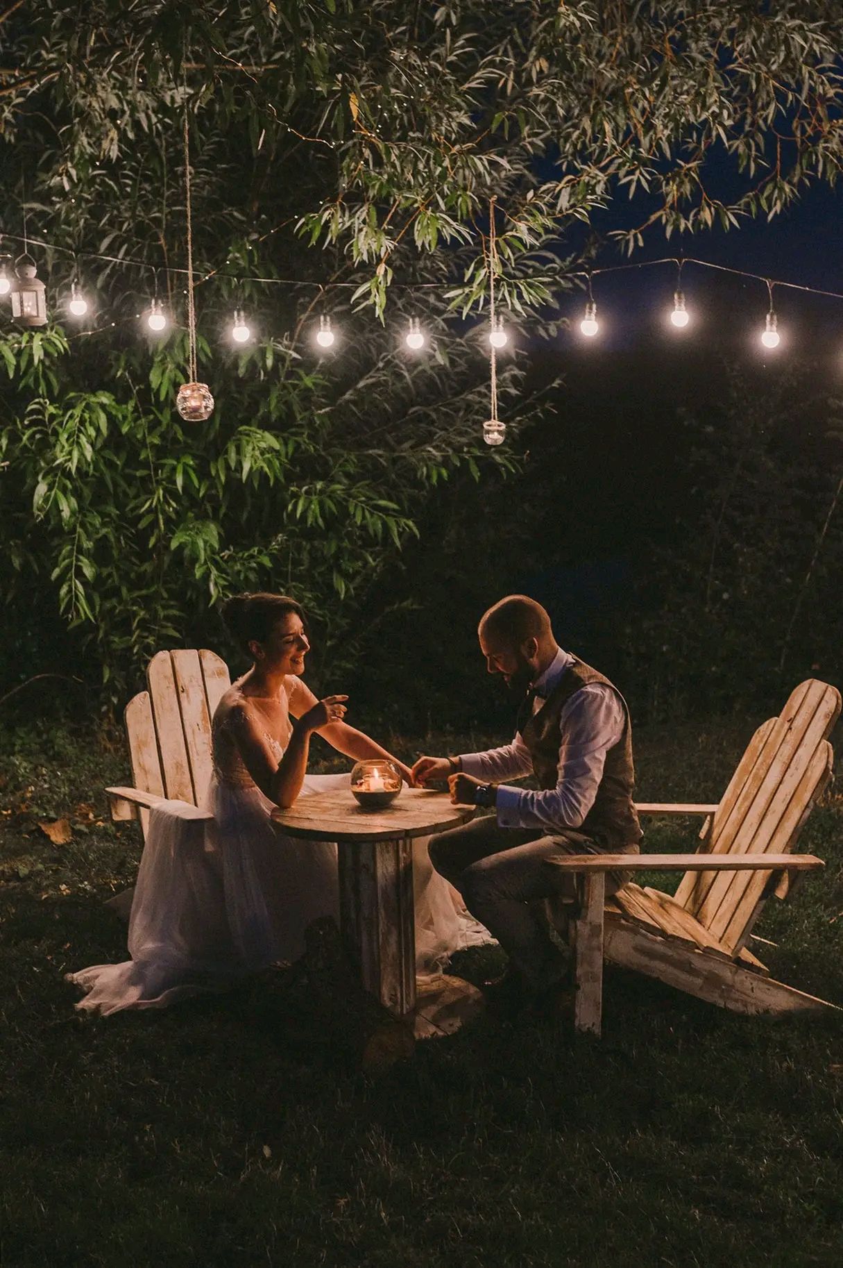A couple enjoys an intimate evening under string lights on Adirondack chairs in a backyard garden setting.