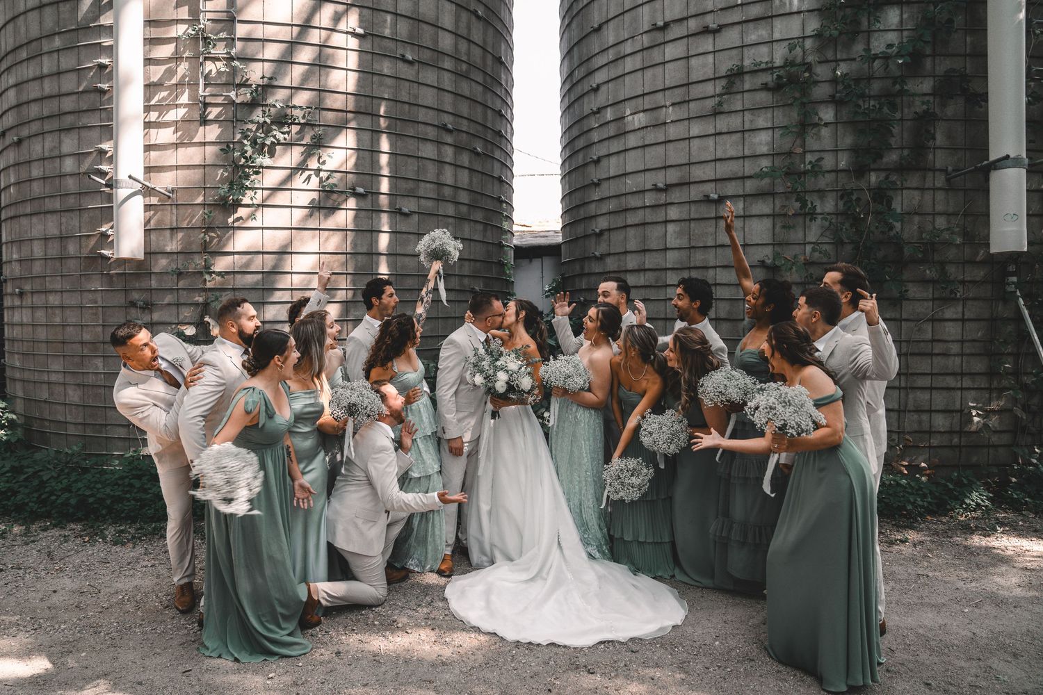 A wedding party in sage green bridesmaid dresses gathers around a bride in white against rustic farm silos.