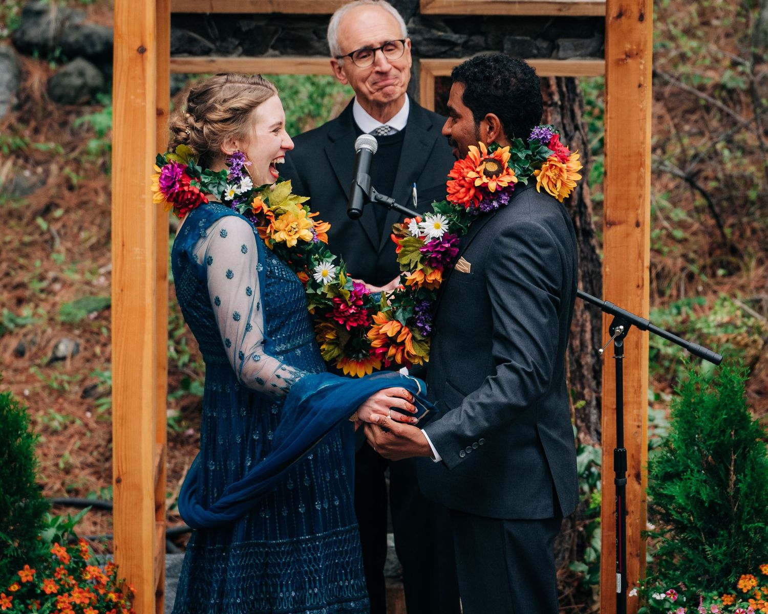 Couple exchanges vows at outdoor multi-cultural wedding ceremony in Leavenworth wearing colorful traditional flower garlands with officiant.