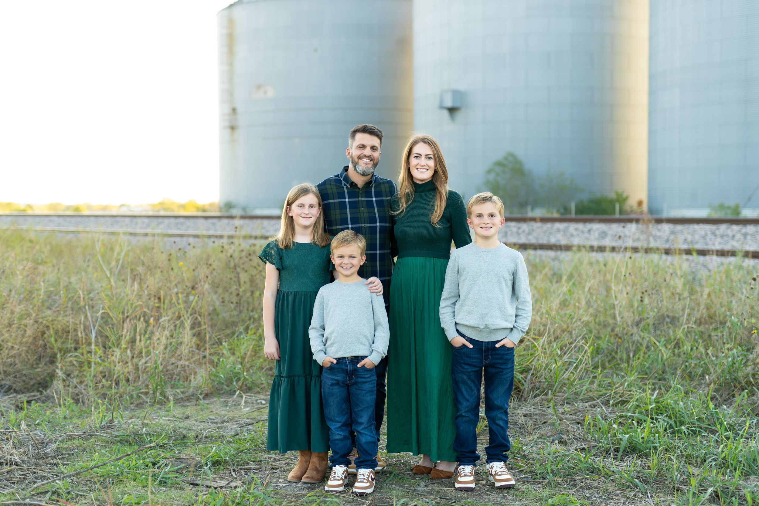 Family of five wearing coordinated green and gray outfits poses together in front of large grain silos at sunset.