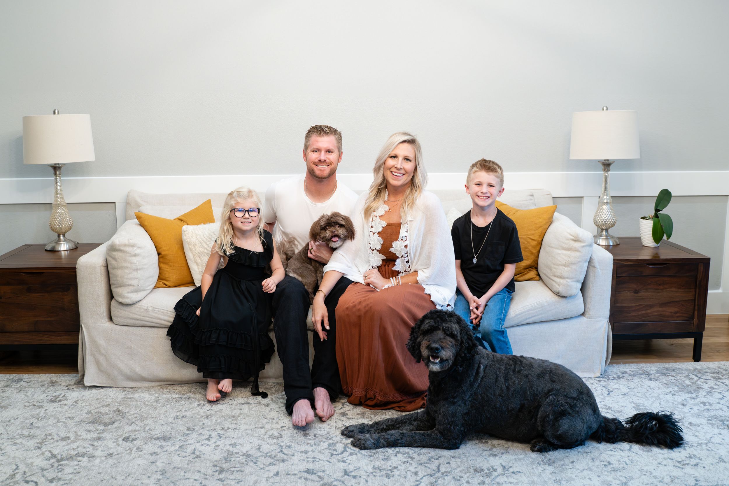 A family relaxes together on a white couch with yellow pillows and two black dogs in a bright living room setting.