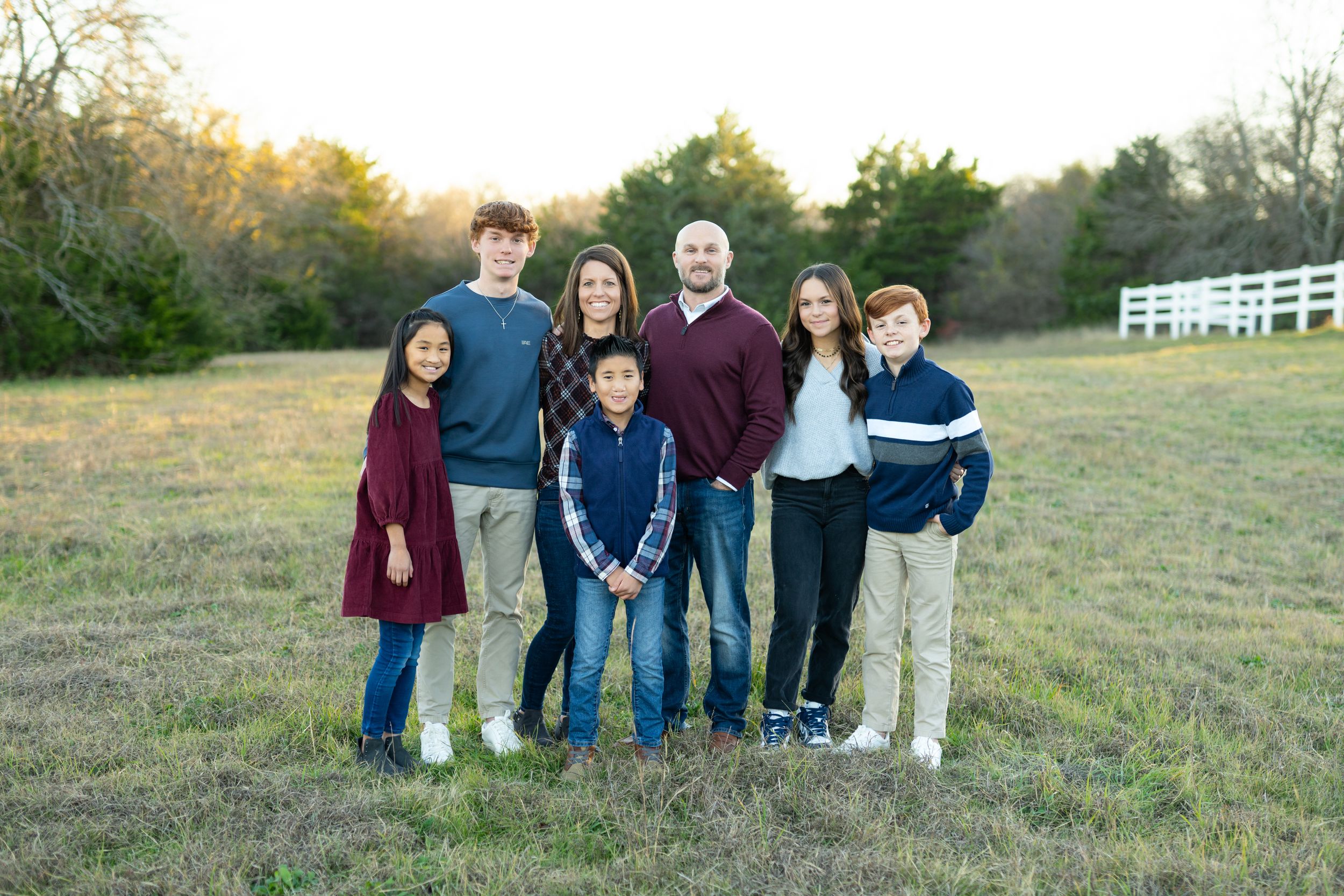 A group stands together in a grassy field at sunset with white fencing visible in the background.