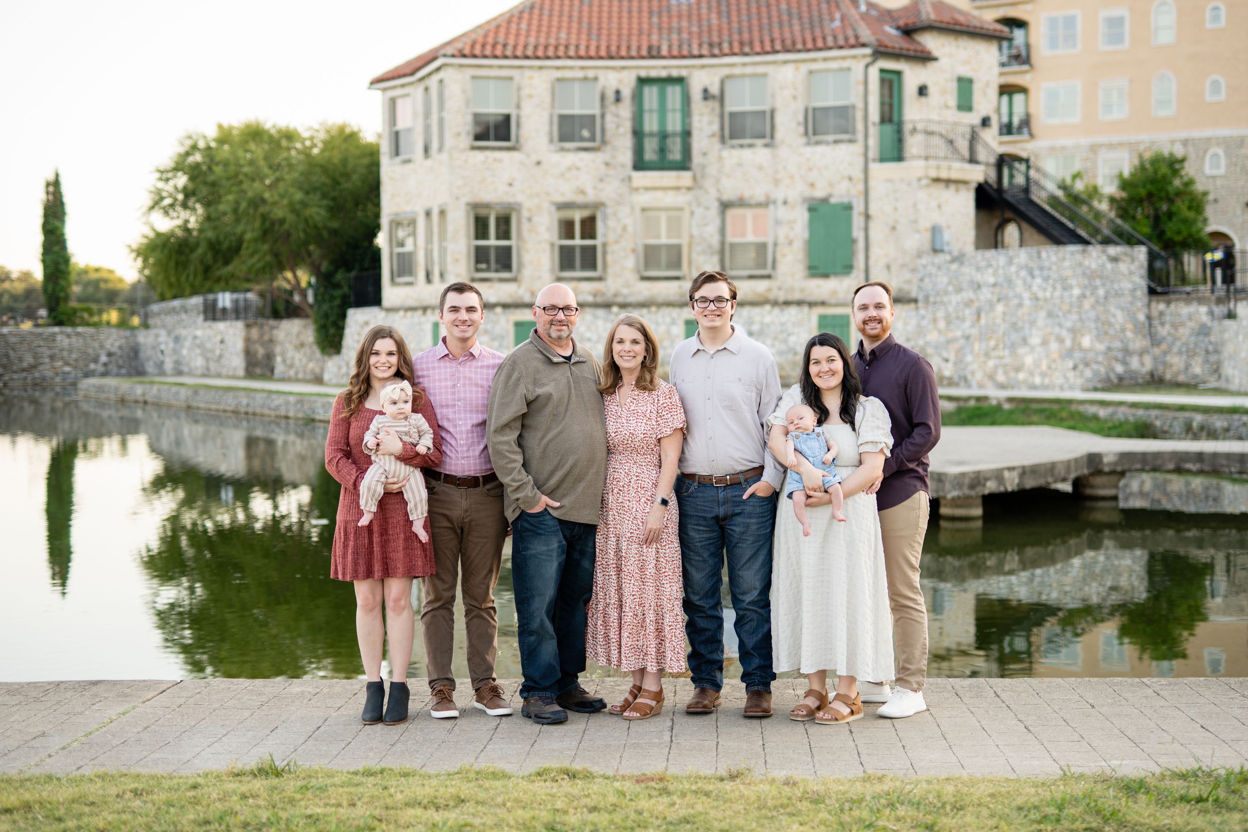 A group poses together by a tranquil lakeside with a white historic building featuring terracotta roof tiles in the backdrop.