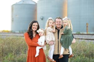 A family poses together in front of large grain silos on a farm during golden hour.