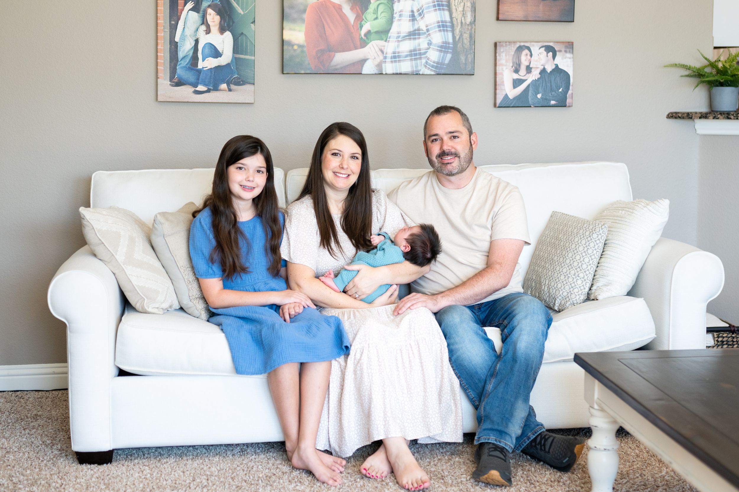 Family sits together on white couch in living room with framed photos displayed on wall behind them.