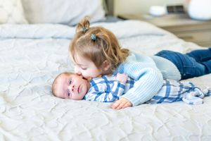 Two children lying together on a white quilted bed, with one wearing a blue and white plaid shirt.