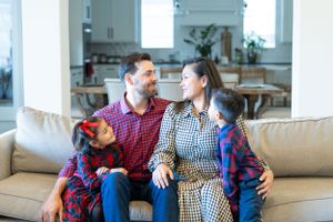 Family relaxing together on beige couch in bright living room with kitchen visible in background.