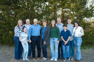 A group of people in casual clothing standing together outdoors against a backdrop of evergreen trees.