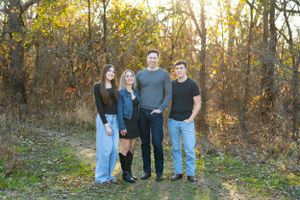 Four people in casual attire stand together on a wooded path during golden hour with autumn foliage in background.