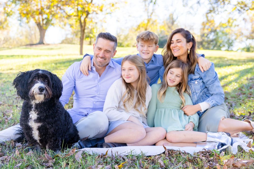 A black dog sits with a family enjoying a picnic on a blanket under autumn trees in a Raleigh park family photo session.