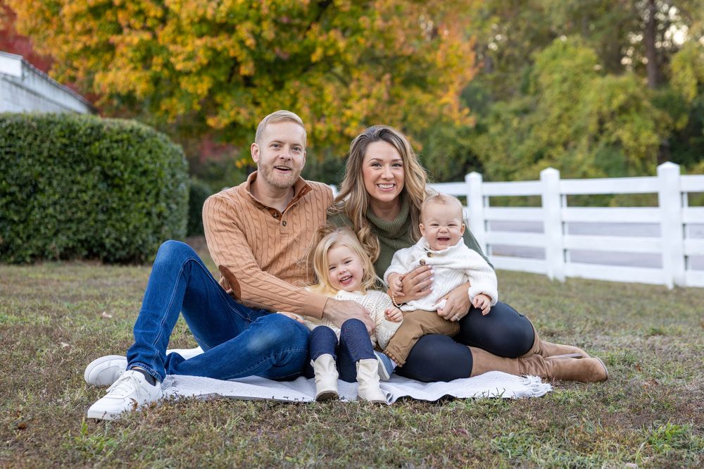 Family sits together on blanket for autumn portrait with white fence and fall foliage in background at Historic Oak View Park in Raleigh.