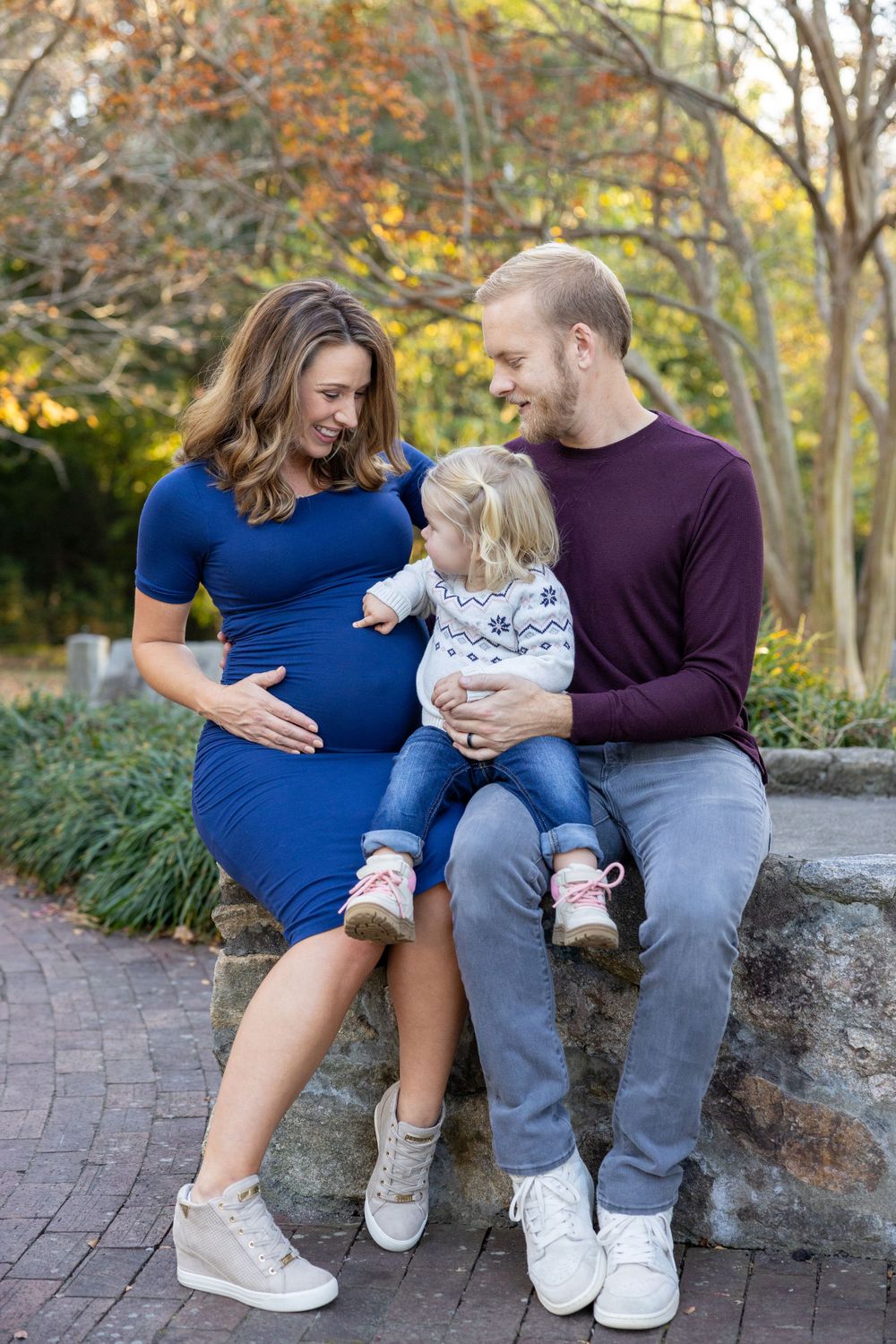 Expecting family sitting on brick path in autumn garden with toddler and woman in blue dress showing baby bump in Raleigh, NC