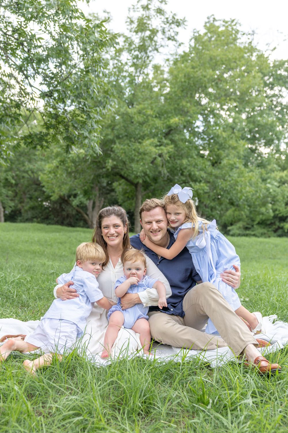 Happy family of five posing on a blanket in a lush green park, parents with three young children in coordinating blue outfits.