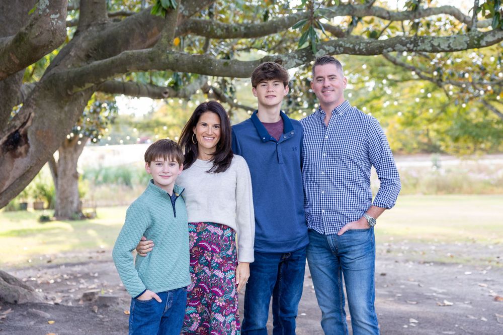 A family of four standing together under a large tree branch at the House on Barber Mill in Clayton, NC.