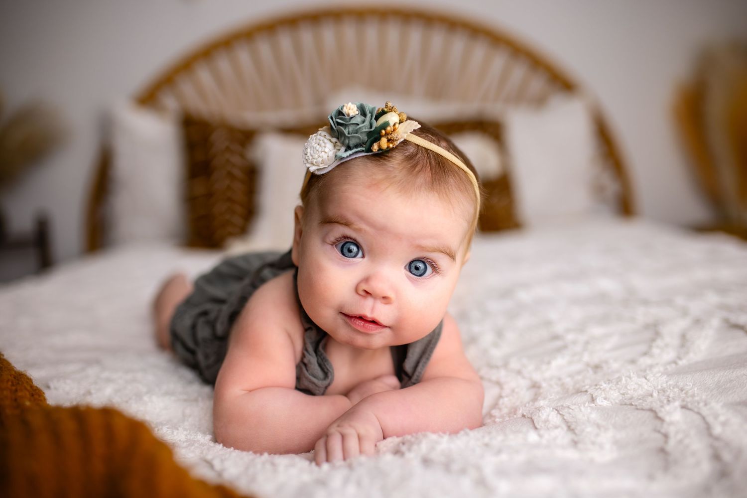 Baby girl with big blue eyes laying on a bed in a photography studio with an adorable green romper and headband and a sweet smile