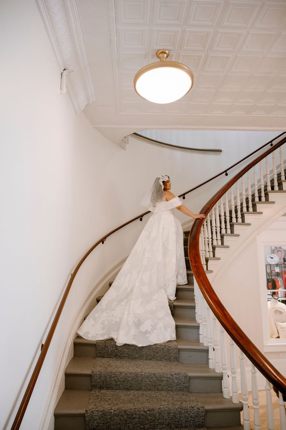 An elegant white wedding dress with a long train displayed on a curved staircase with white walls and wooden railings.