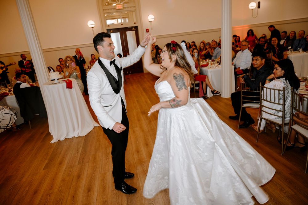 A couple performs their first dance at their wedding reception in a ballroom with hardwood floors and guest tables.