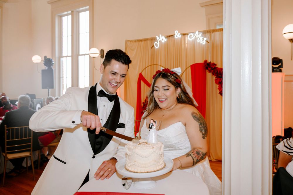 A bride and groom smile together while cutting their wedding cake at an indoor reception venue.