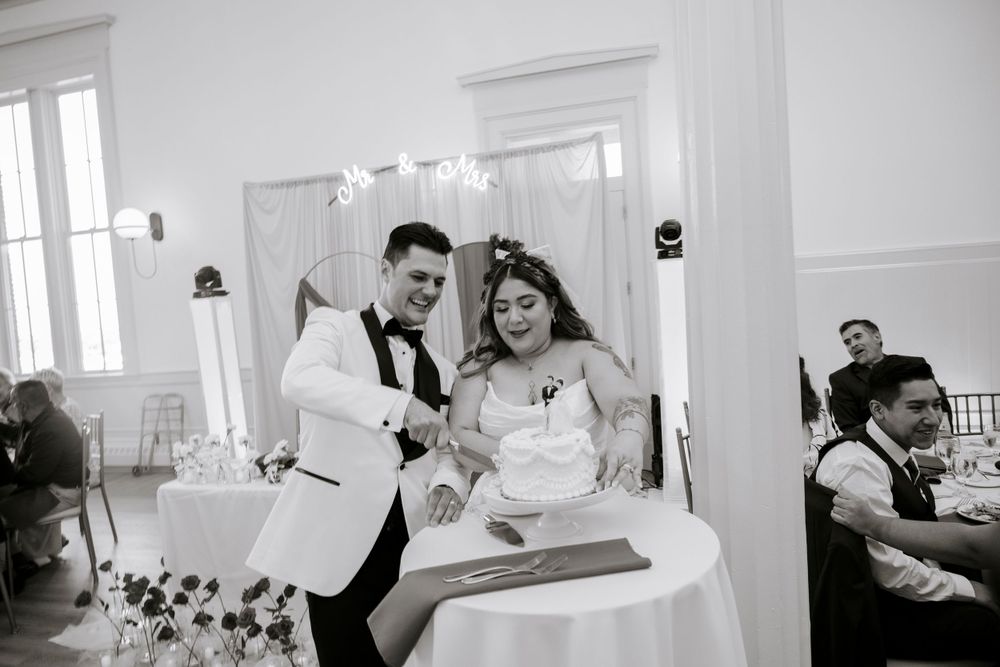 A black and white photo captures a joyful cake-cutting moment at an intimate wedding reception.