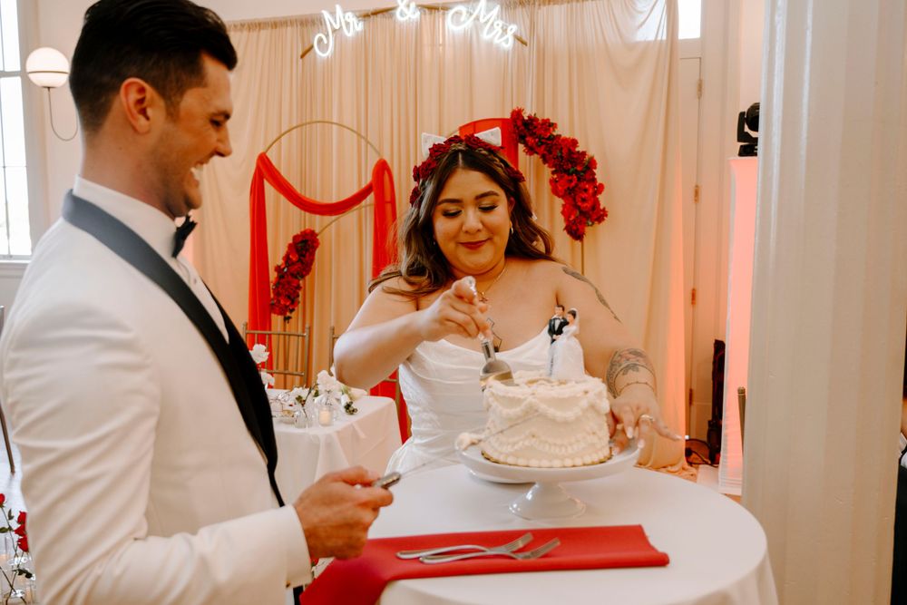 Newlyweds in white attire share a cake-cutting moment at their reception with red decor accents.