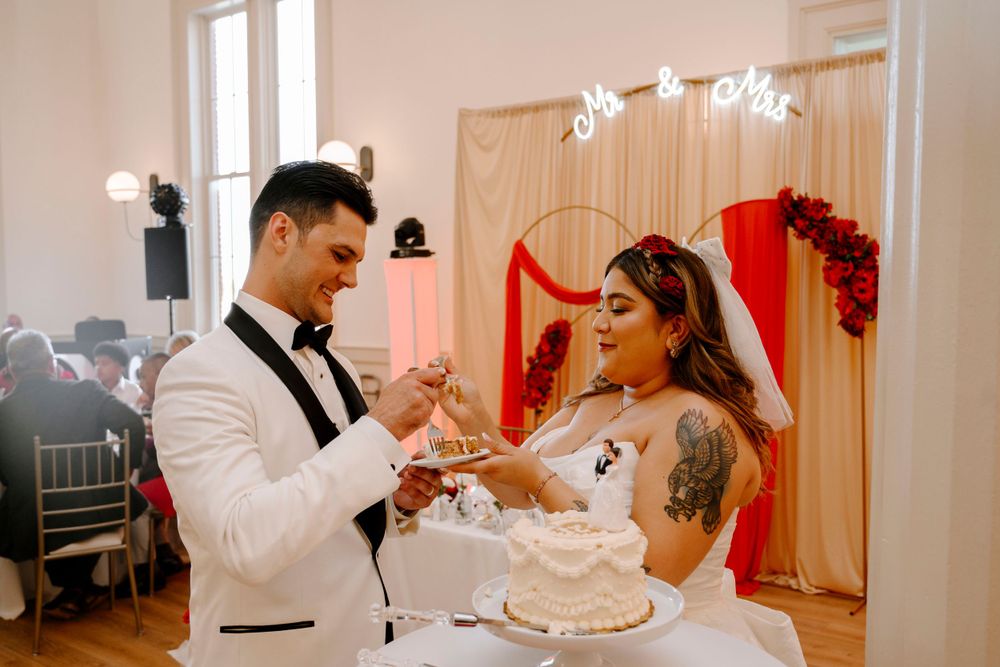 Newlyweds share a playful moment while cutting their white wedding cake in front of a decorated arch.