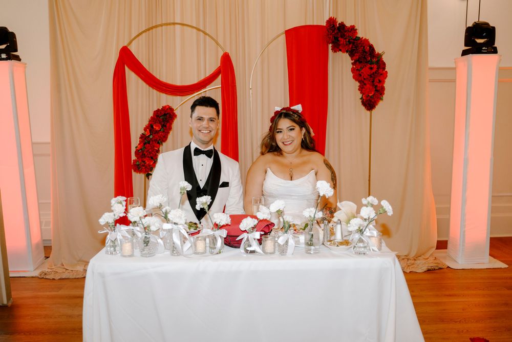 A wedding reception setup with a decorated sweetheart table featuring red floral arrangements and white decor elements.