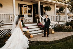Bride in a white gown surprises groom on the steps of a charming farmhouse decorated for fall.