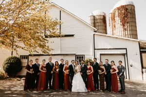 Wedding party posing in front of a white barn with silos, bridesmaids in rust red dresses.