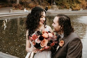 Bride with dark curly hair holds autumn bouquet while smiling at groom near a Central Park fountain.
