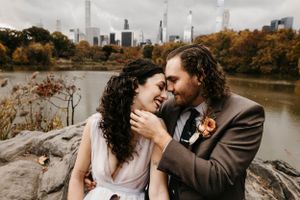 Bride and groom share a tender moment on rocks with the Manhattan skyline in the autumn background.