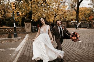 Laughing bride in flowing gown and groom walk hand in hand on a Central Park path in autumn.