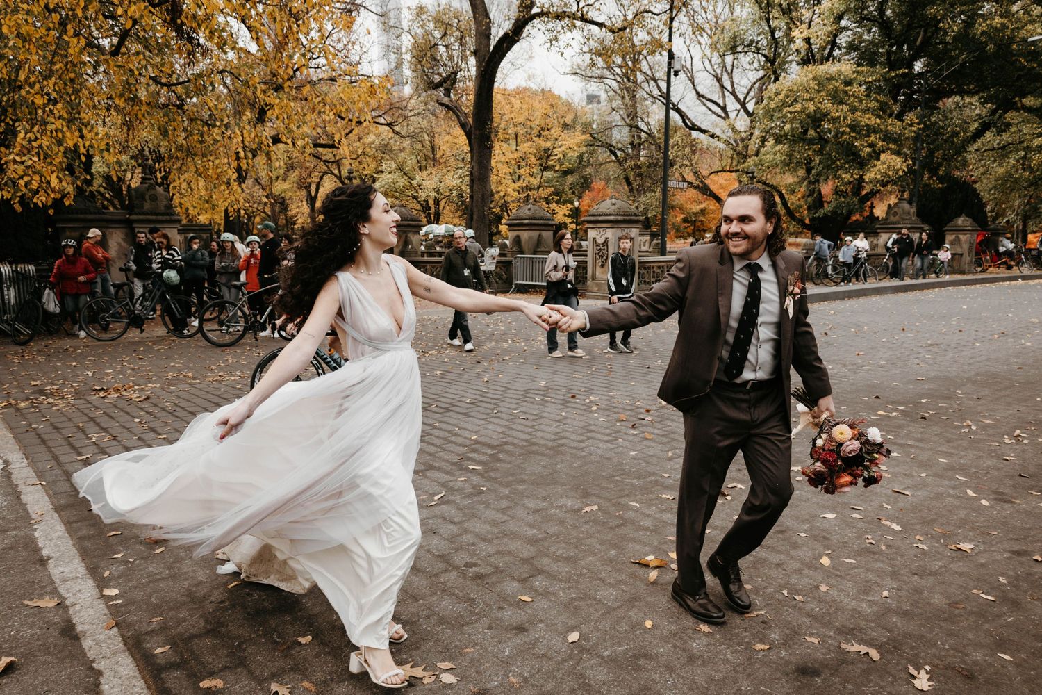 Joyful bride and groom dance together on a Central Park cobblestone path surrounded by fall foliage.