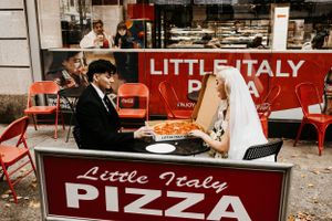 Newlyweds share a pizza slice at an outdoor table at Little Italy Pizza on their wedding day.