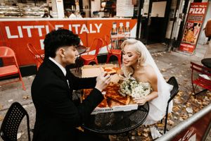Bride holding bouquet and groom share a pizza slice at Little Italy Pizza on their wedding day.
