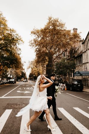 Bride and groom cross a New York City street in wedding attire surrounded by autumn foliage.