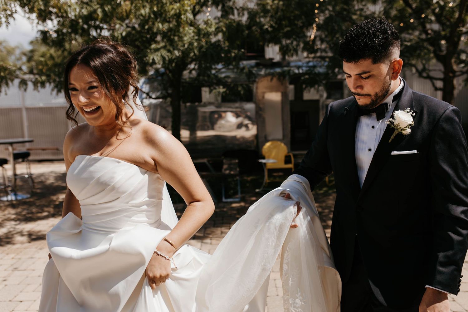 Bride laughing in white strapless gown lifts dress while groom in black tuxedo assists outdoors.