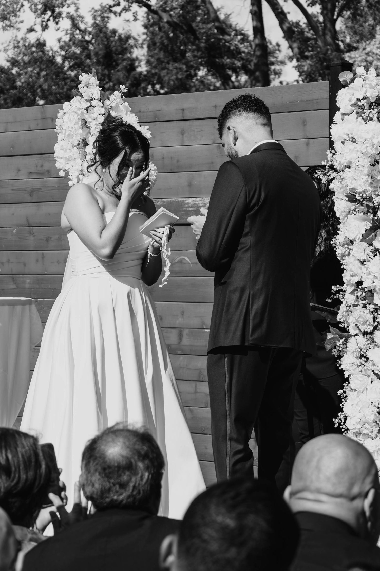 Emotional bride reads vows while groom listens at floral arch in black and white wedding photo.