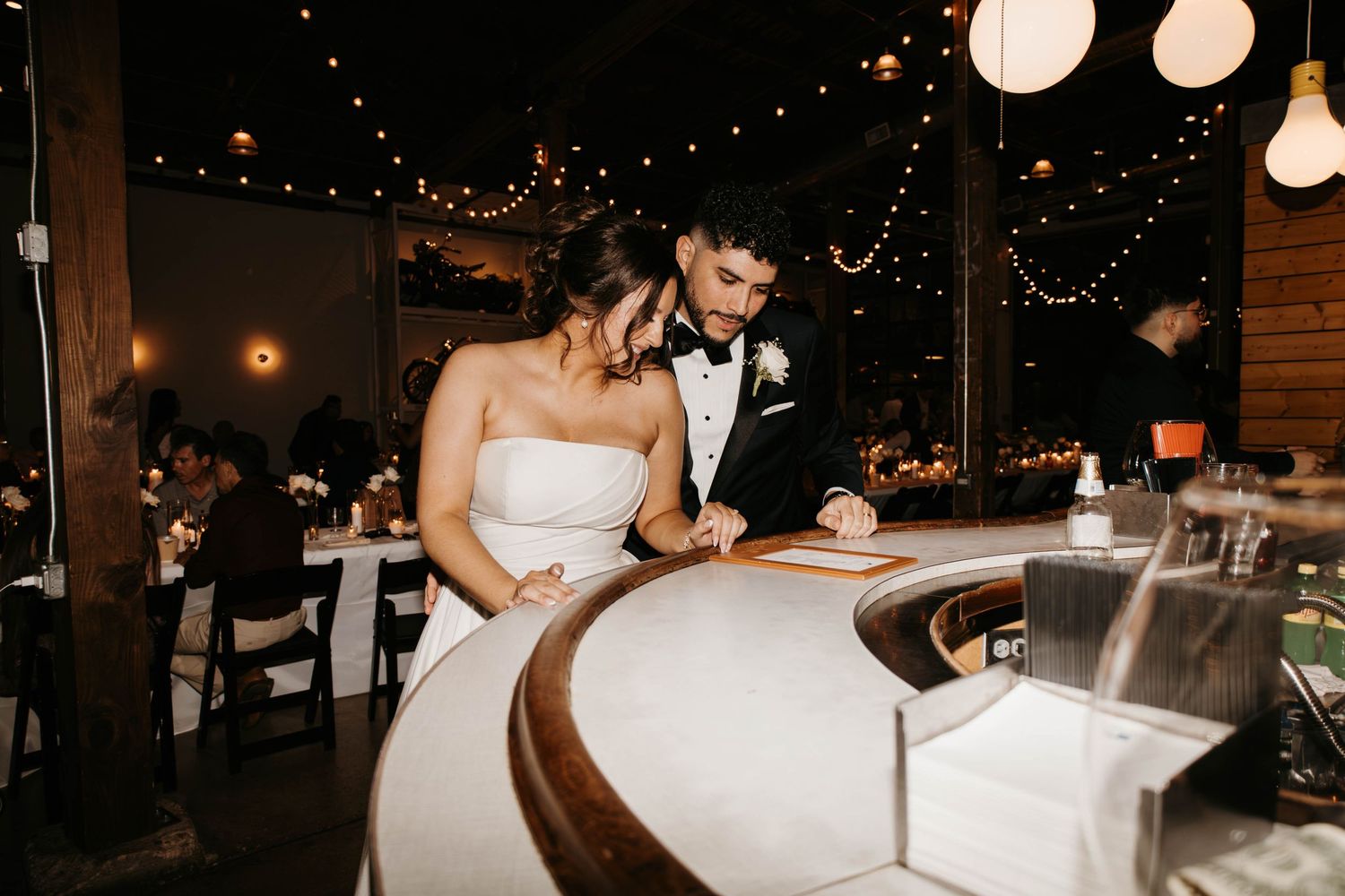 Bride and groom sign wedding documents at candlelit reception venue with string lights overhead.