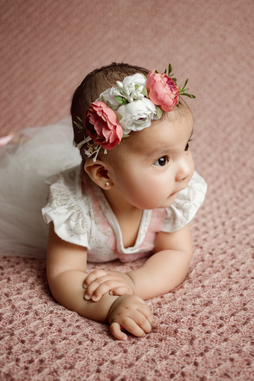 A baby in a white lace dress lies on pink carpet wearing a floral headband with pink and white roses.