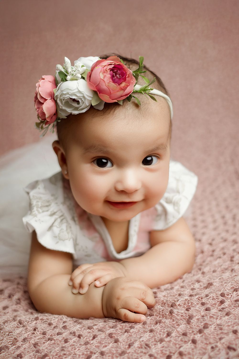 A baby wearing a delicate lace dress and floral crown with pink and white roses lies on a dusty pink textured backdrop.